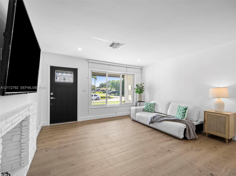 Living space featuring wood-finish flooring, a painted brick fireplace, a black front door with an integrated window, and a large picture window