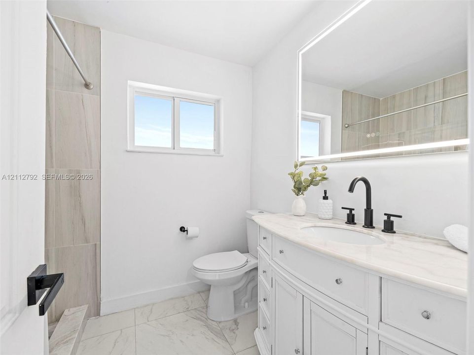 Bathroom featuring a vanity with stone countertop, integrated sink, matte black widespread faucet, LED-backlit mirror, and neutral tile shower surround