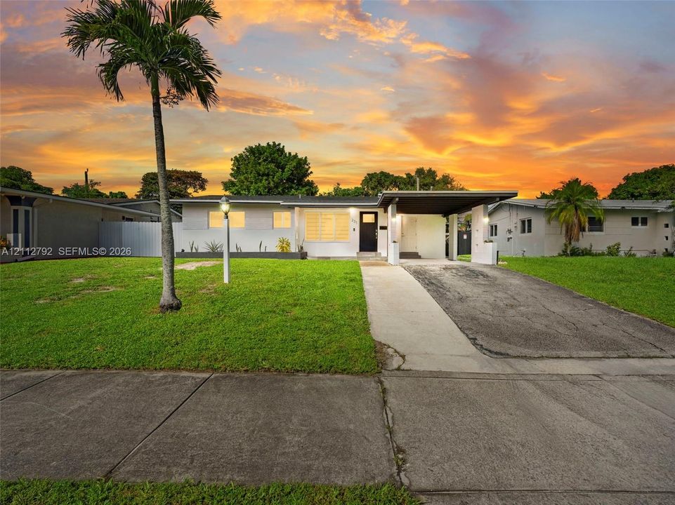 Single-story exterior featuring a painted masonry facade, an integrated carport, a concrete driveway, and mature palm landscaping