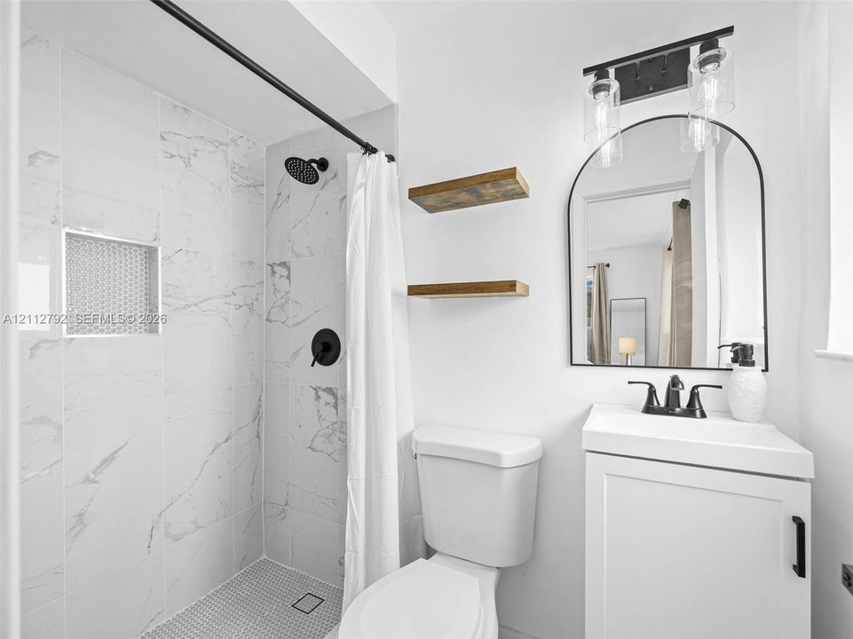 Bathroom featuring a shower stall with stone-finish tiling, a built-in recessed shower niche, a white vanity cabinet, a matte black arched mirror, and wood-finish wall-mounted shelving