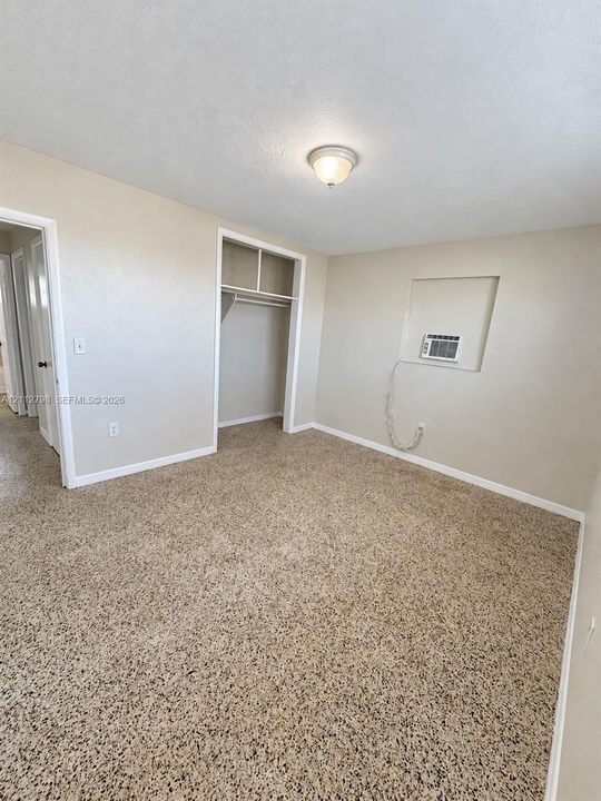 Interior space featuring neutral wall tones, wall-to-wall textured carpeting, a recessed closet with an integrated shelf and rod, and a built-in wall-mounted climate control unit
