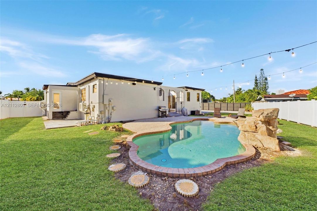 Backyard featuring an in-ground swimming pool with a stone rock feature, concrete patio, perimeter vinyl fencing, and overhead string lighting
