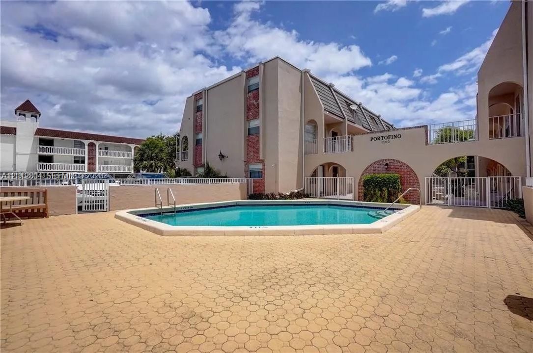 Exterior common area featuring an inground swimming pool with metal ladder access, surrounded by a paved deck and enclosed by a white security fence