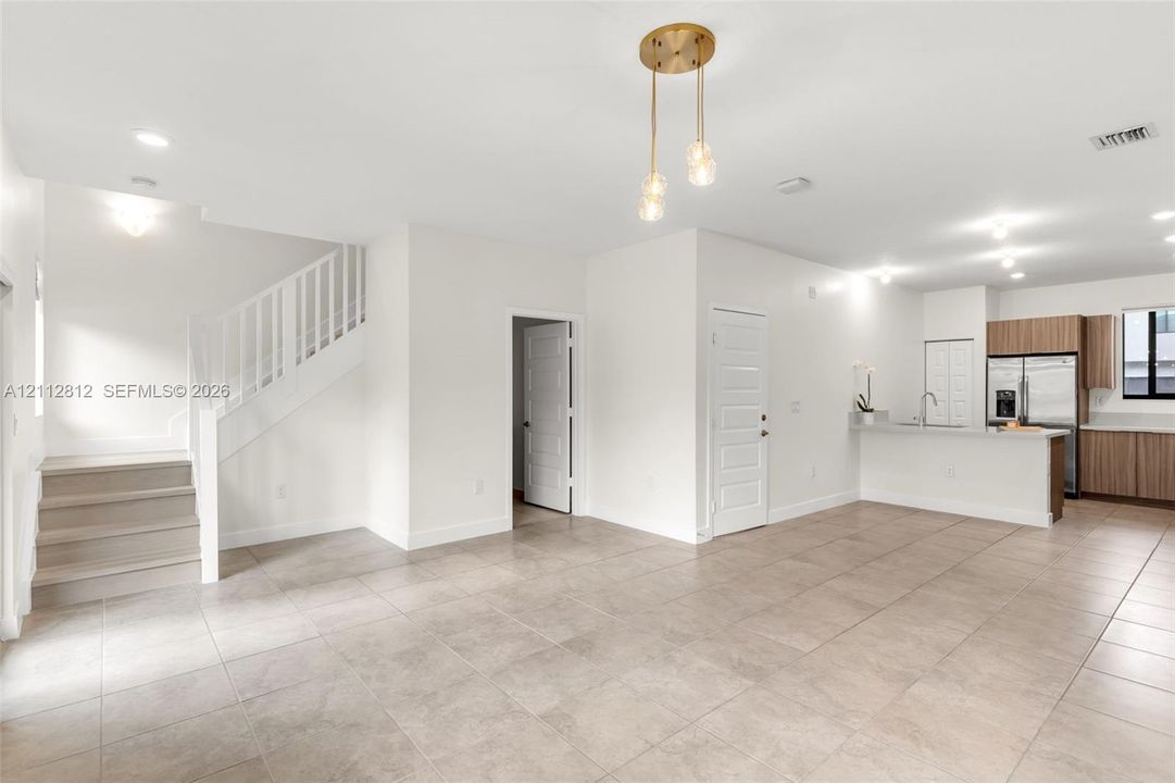 Open-concept interior featuring large-format neutral tile flooring, a staircase with a white railing, a contemporary three-globe pendant light fixture, and wood-finish cabinetry