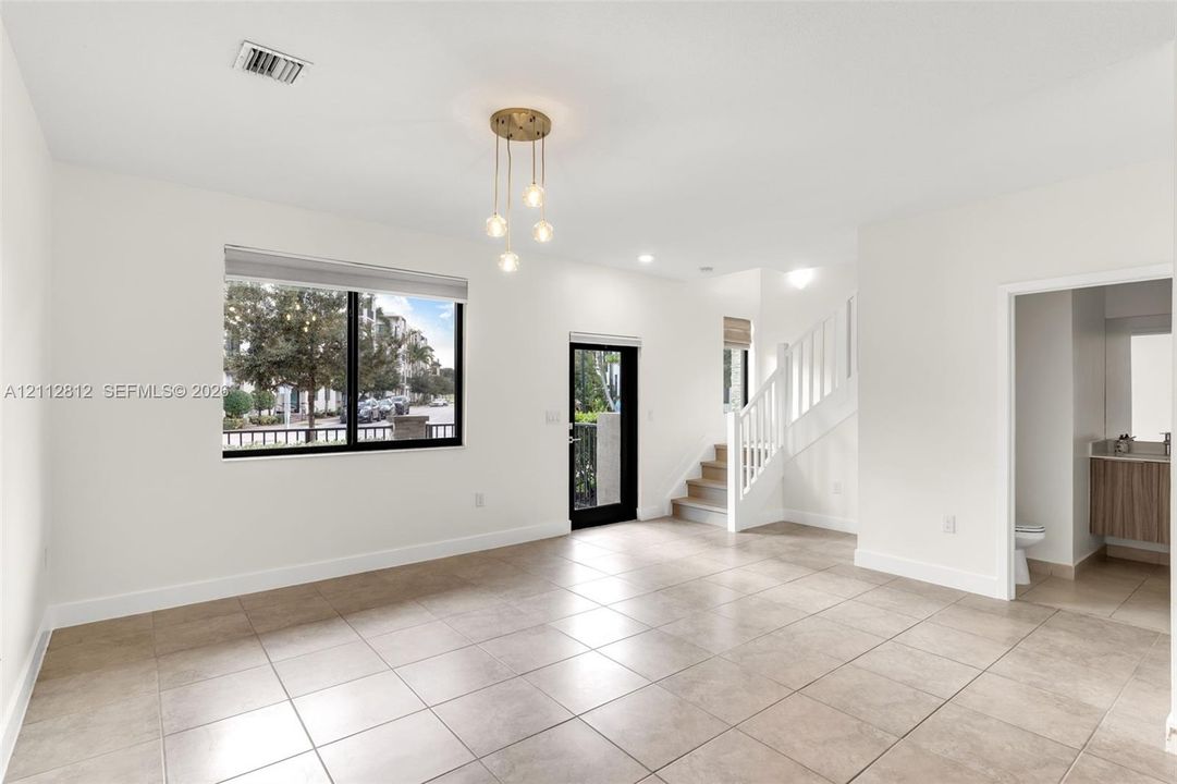 Open-concept interior featuring large-format tile flooring, multi-light gold-finish pendant fixture, black-framed picture window, and a staircase with white balusters