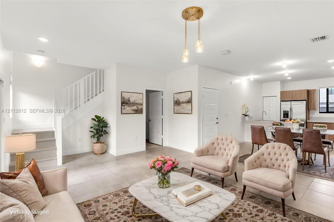 Open-concept interior featuring neutral tile flooring, staircase with white railing, recessed lighting, multi-pendant brass light fixture, and kitchen cabinetry with wood-finish accents