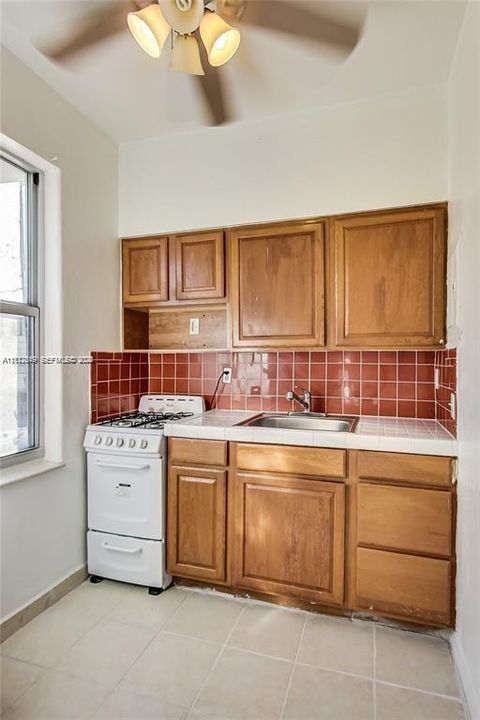 Kitchen featuring wood cabinetry, white tiled countertops, a red tile backsplash, a gas range, and a stainless steel sink with a mounted faucet