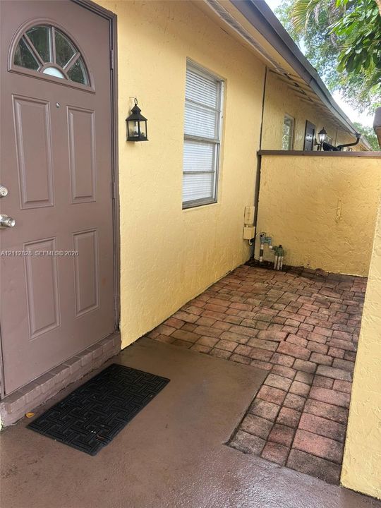 Exterior entryway featuring a solid-paneled door with an arched glass insert, exterior lantern-style wall lighting, and brick paver flooring