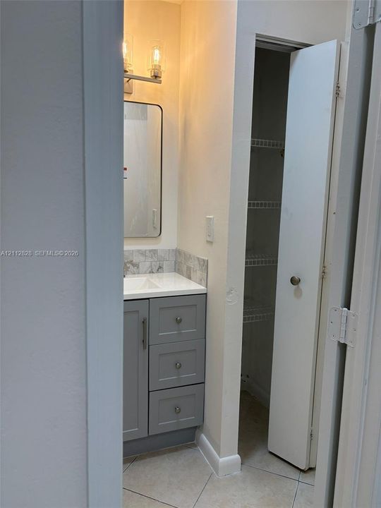 Gray vanity cabinet with a white countertop, tiled backsplash, rectangular mirror with metallic frame, two-light fixture, and built-in closet with wire shelving