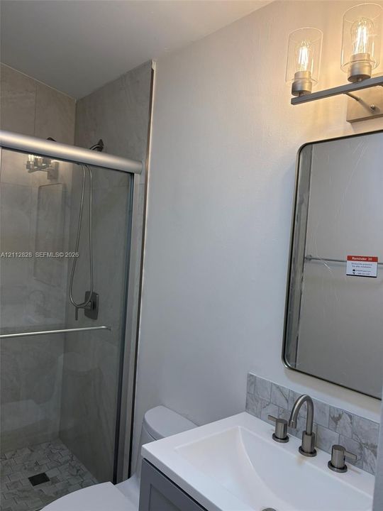 Shower with sliding glass doors and tiled surround, vanity with stone-style backsplash, integrated sink, metal wall-mounted light fixture, and framed mirror