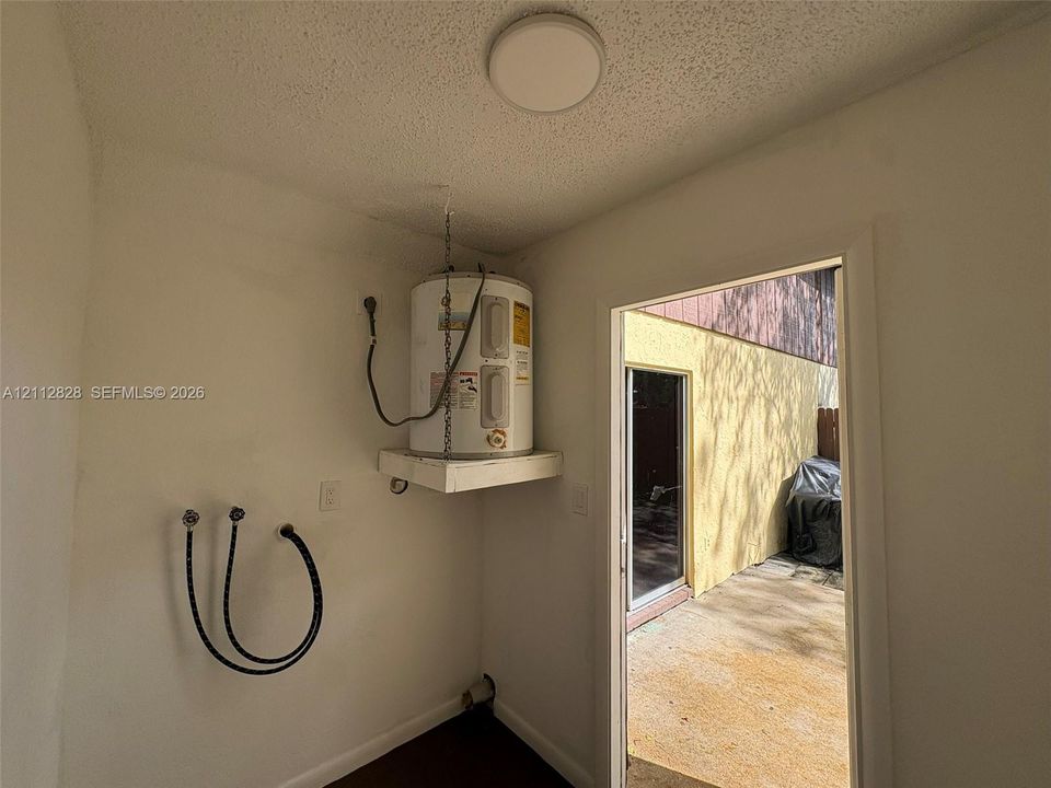 Utility space featuring a wall-mounted water heater, dedicated plumbing hookups, and a flush-mount ceiling light fixture, with access to an exterior concrete patio