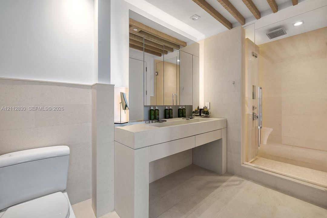 Stone-surfaced floating vanity, ceiling with exposed wood-finish beams, integrated medicine cabinet, and glass-enclosed walk-in shower with stone tile wall surfacing