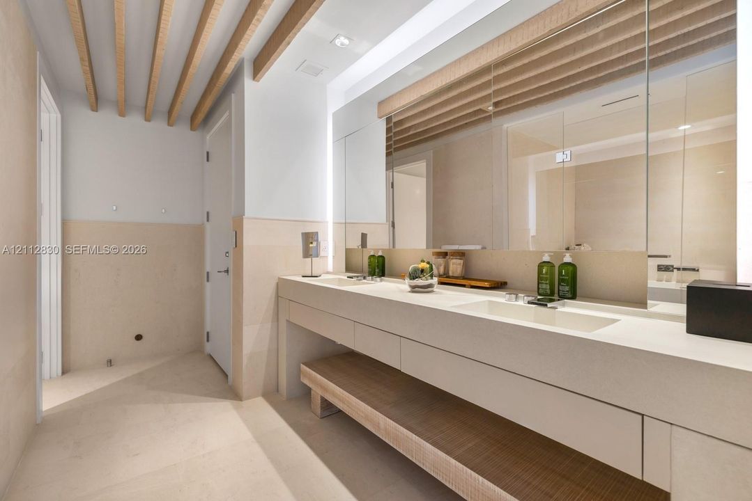 Dual vanity with integrated sinks, stone-surface countertop, full-width mirror with integrated lighting, exposed wood ceiling beams, and stone-finish wall cladding