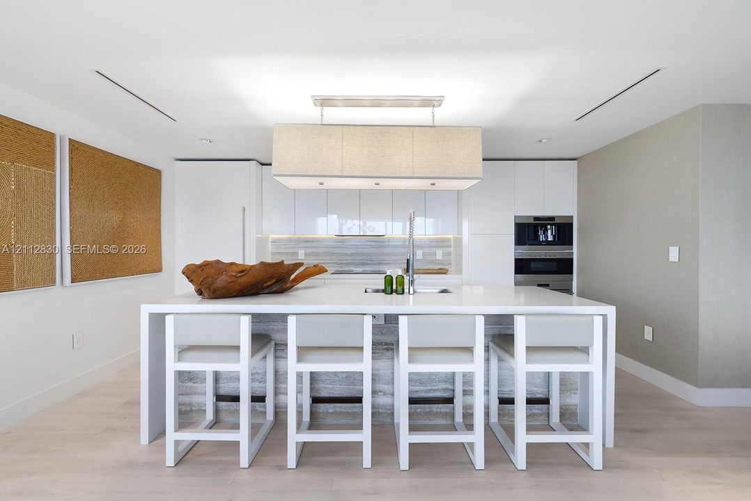 Kitchen featuring high-gloss cabinetry, stone-finish island waterfall countertop, integrated stainless steel appliances, and rectangular pendant light fixture over a breakfast bar