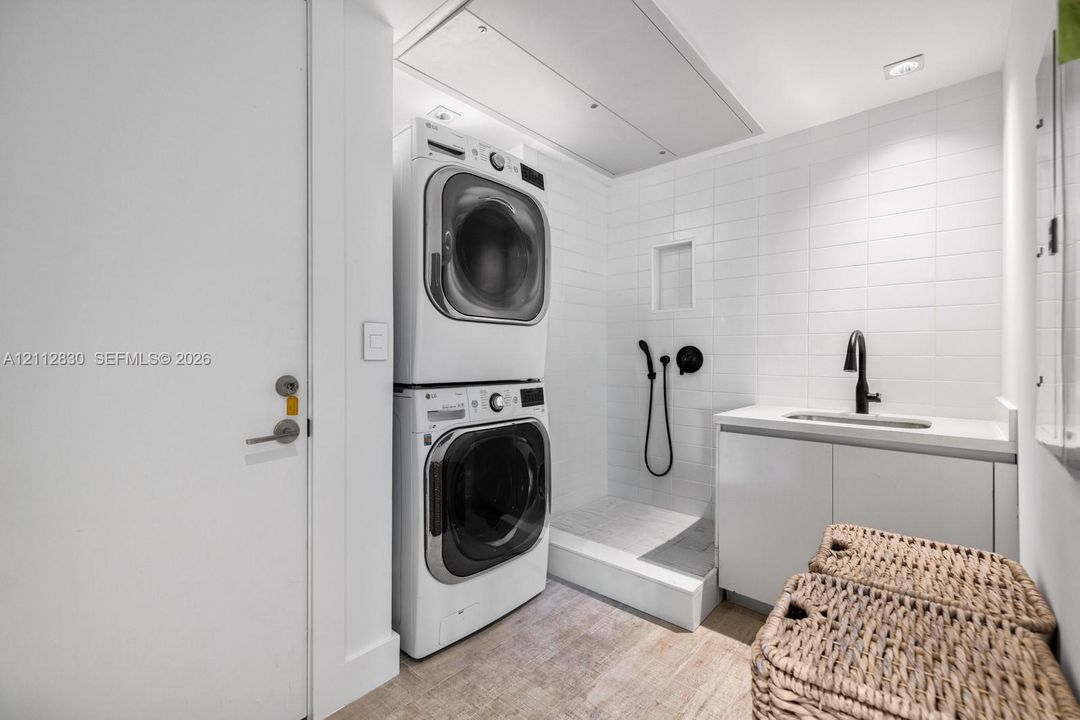 Laundry area featuring stacked appliances, tiled shower enclosure with handheld sprayer, built-in cabinetry, solid-surface countertop, and matte black faucet