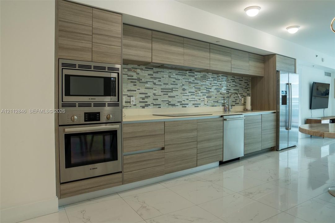 Kitchen featuring flat-panel wood-finish cabinetry, stainless steel appliances, multi-tonal glass tile backsplash, stone-finish countertops, and polished large-format flooring