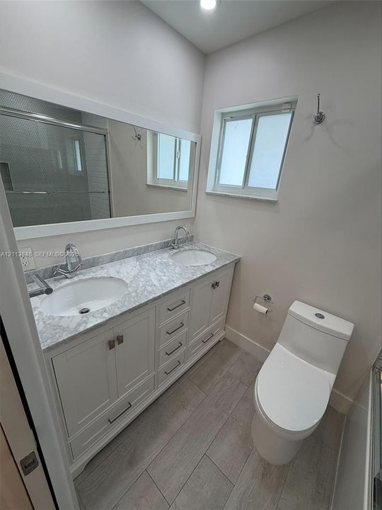 Dual-sink vanity with stone-finish countertop, white cabinetry with brushed metal hardware, wall-to-wall mirror, wood-finish flooring, and a low-profile elongated toilet