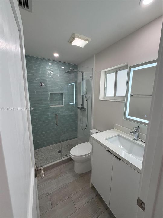 Walk-in shower with floor-to-ceiling glass paneling, blue subway tile surround, and integrated recessed shelving, featuring wood-finish tile flooring, a white vanity with integrated basin, and a wall-mounted LED-backlit mirror