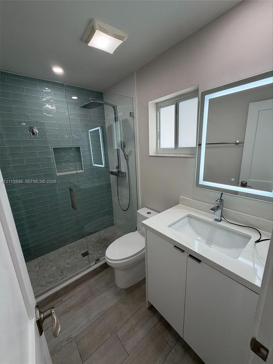 Bathroom featuring a glass-enclosed walk-in shower with blue subway tile, a built-in shower niche, a white vanity with a quartz countertop, a lit mirror, and wood-finish flooring