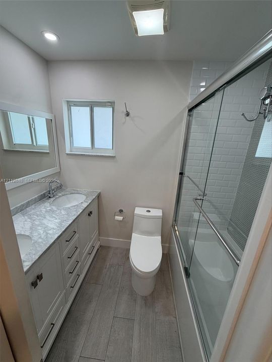 Bathroom featuring a dual-sink vanity with stone-finish countertop, wood-finish tile flooring, and a glass-enclosed shower with subway tile surround
