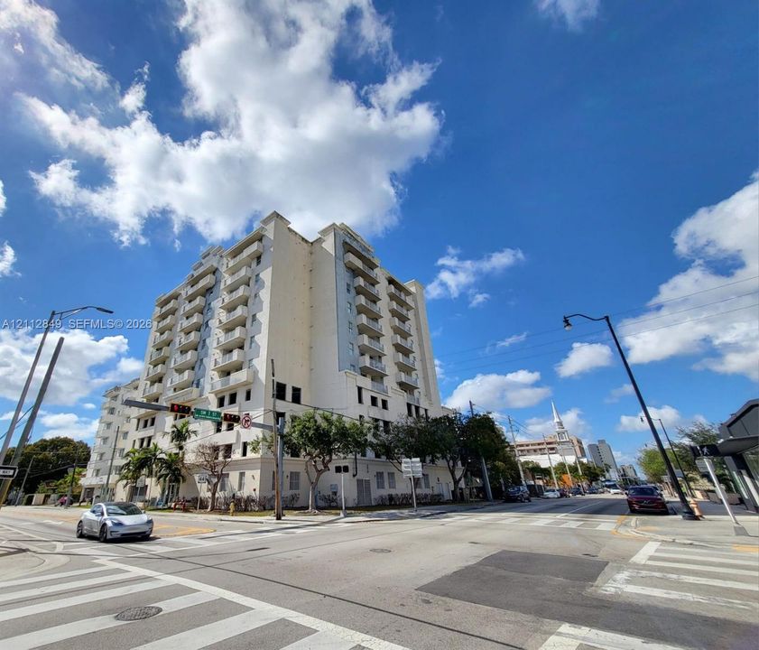 High-rise multi-story building featuring a white facade, stacked private balconies, ground-level parking structure, and mature landscaping