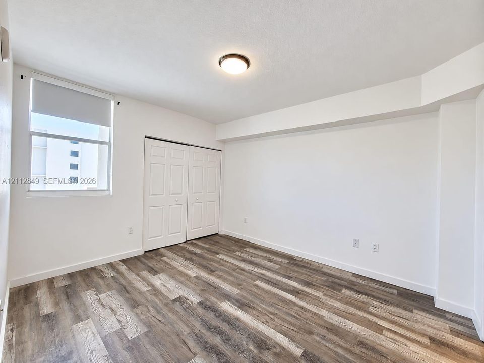 Interior room featuring wood-finish flooring, a vertical window with a roller shade, a dual-panel closet door, and a flush-mount ceiling fixture
