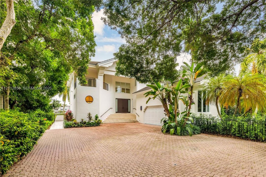Stucco exterior featuring a circular accent window, elevated entryway with stone steps, multi-car garage, and a spacious paved driveway shaded by mature trees