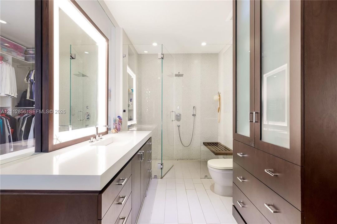 Custom dark-wood cabinetry, white composite countertops, integrated LED-backlit mirror, glass-enclosed walk-in shower with pebble-tiled wall, and built-in floor-to-ceiling storage