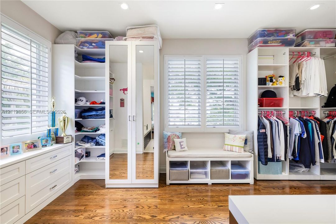 Custom closet featuring white built-in cabinetry, floor-to-ceiling mirrored doors, integrated window seating, plantation shutters, and wood-finish flooring