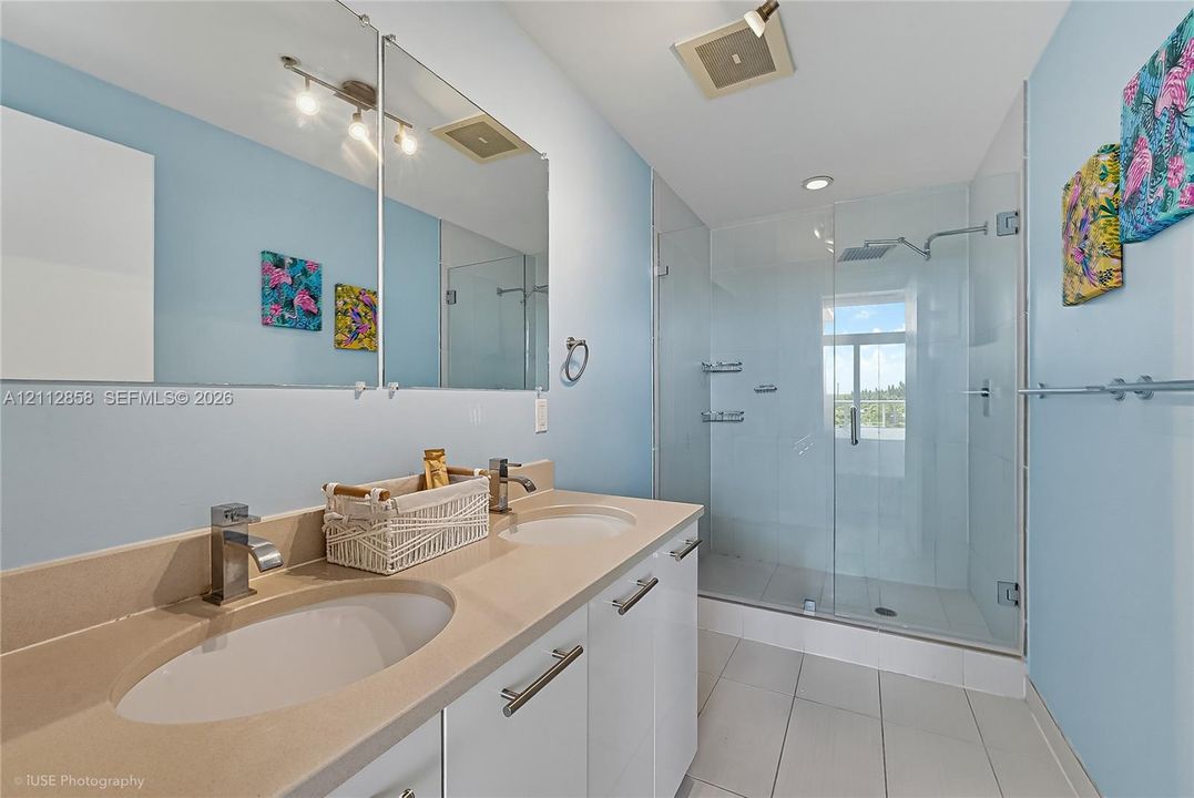 Dual-sink vanity with a solid-surface countertop, full-width mirror, glass-enclosed shower with a rainfall showerhead, and tile flooring