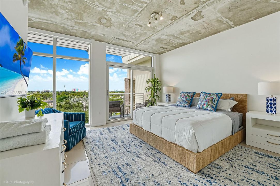 Exposed concrete ceiling, floor-to-ceiling windows, sliding glass door access to a balcony, and large-format tile flooring