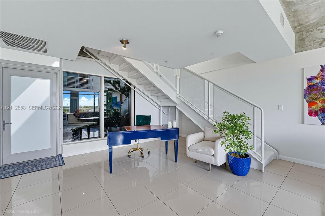 Open-concept interior featuring large-format tile flooring, exposed concrete ceiling, industrial-style staircase with glass railing, and floor-to-ceiling windows