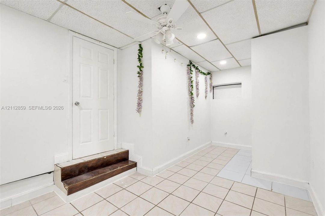 Tiled flooring, drop ceiling with recessed lighting, integrated ceiling fan, and wood-finish step leading to a six-panel door