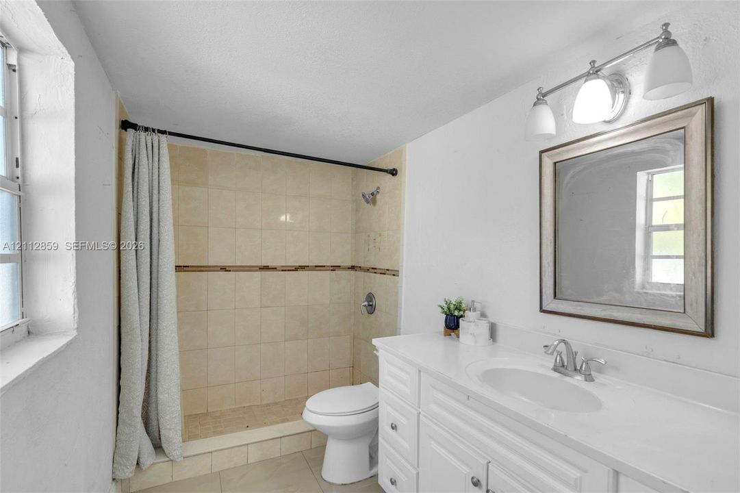 Bathroom featuring a white vanity with an integrated sink, a rectangular framed mirror, a triple-light fixture, and a tiled shower stall with a decorative accent border