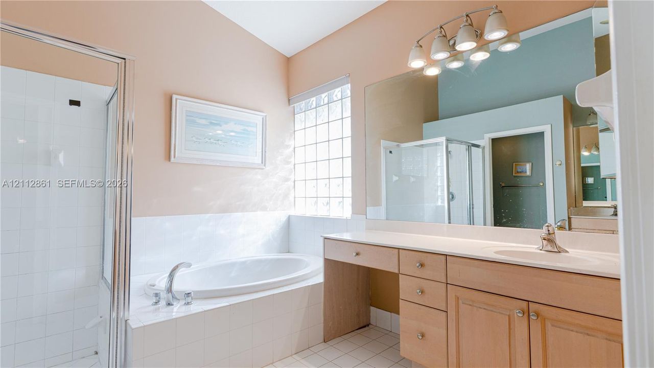 Bathroom featuring a soaking tub with tile surround, glass-enclosed shower, wood-finish vanity with countertop sink, glass block window, and multi-bulb vanity lighting