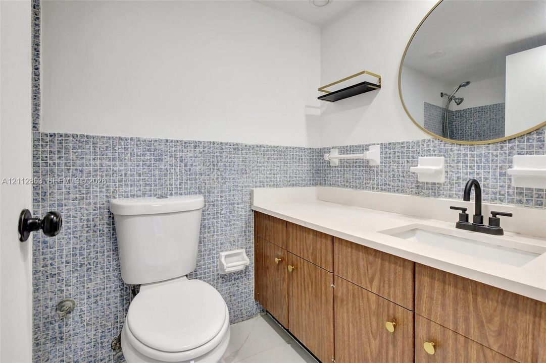 Vanity featuring wood-finish cabinetry, a solid surface countertop with an integrated basin, a matte black faucet, a gold-rimmed oval mirror, and blue mosaic tile wall accents