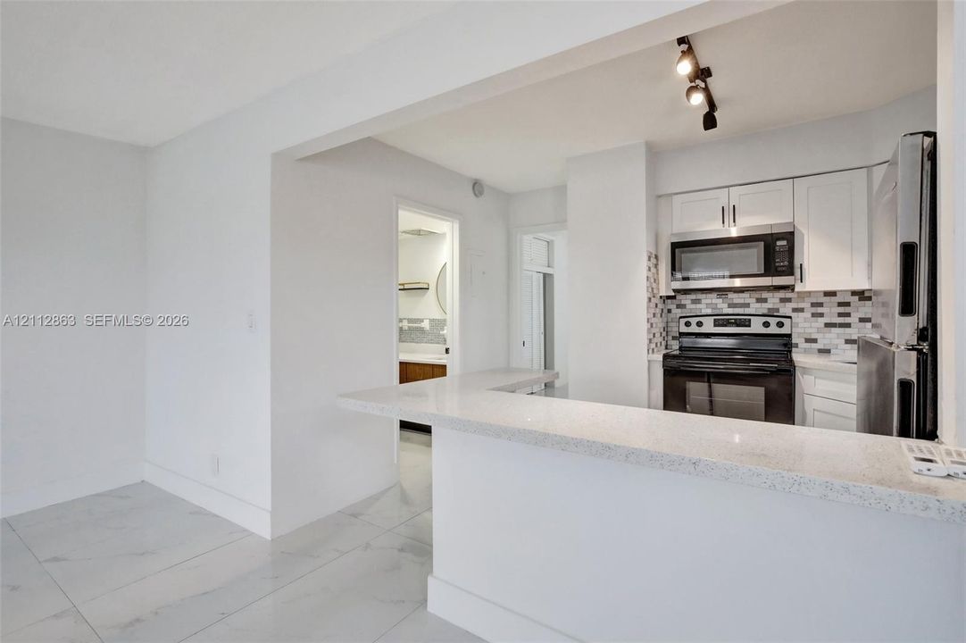 Kitchen featuring white shaker-style cabinetry, a quartz countertop breakfast bar, a stone-tiled backsplash, stainless steel appliances, and large-format porcelain floor tiling