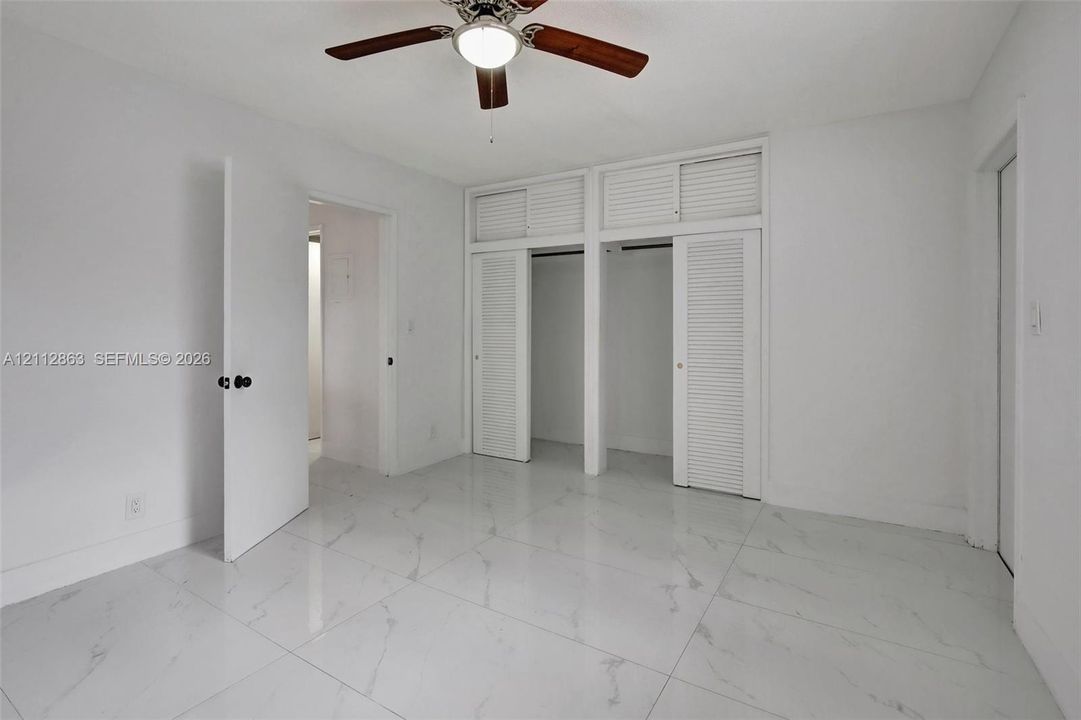 Polished stone-finish flooring, ceiling fan with light fixture, and dual reach-in closets featuring louvered doors with overhead storage compartments