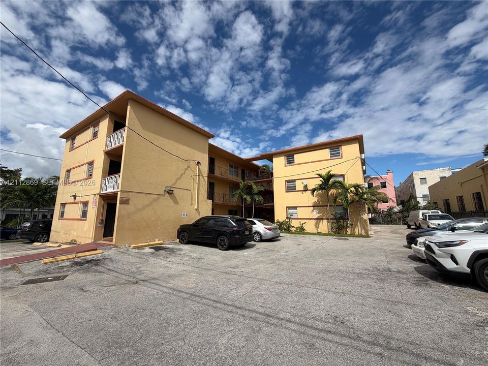 Three-story multi-family exterior featuring a concrete facade, metal railings, and surface parking