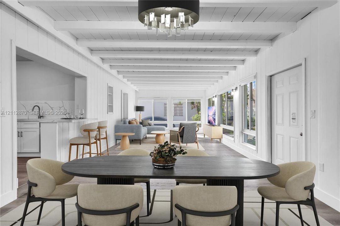 Open-concept area featuring white vertical wood paneling, exposed white ceiling beams, tiered modern chandelier, and multiple large windows