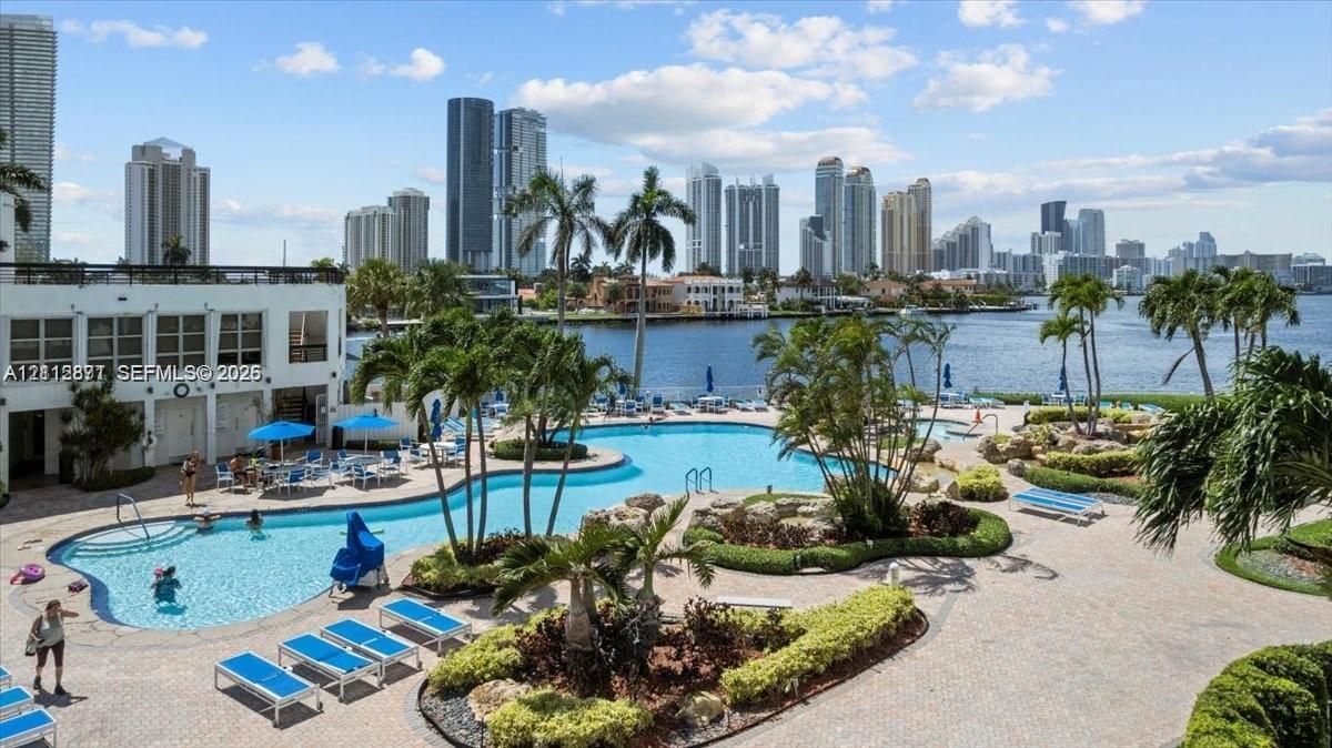 Waterfront pool area featuring a paved sun deck, mature palm trees, decorative landscaping, and a backdrop of the city skyline
