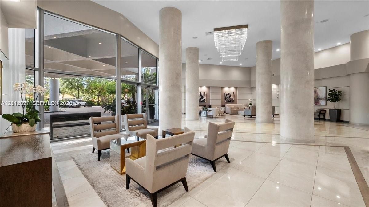 Grand foyer featuring floor-to-ceiling glass paneling, polished stone flooring, structural columns, and tiered crystal chandelier lighting
