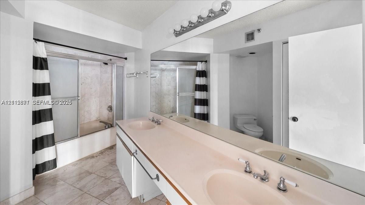 Bathroom featuring a dual-sink vanity with a continuous countertop, expansive wall-mounted mirror, multi-bulb overhead lighting, and a bathtub-shower combination with sliding glass doors