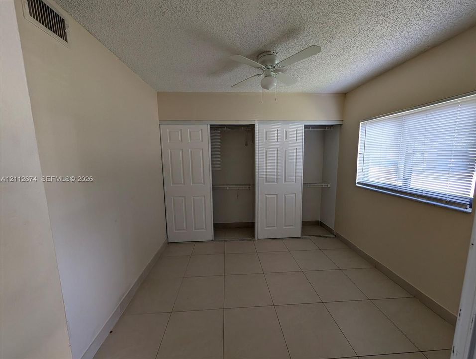 Neutral tile flooring, dual-door reach-in closets, window with horizontal blinds, and a ceiling fan