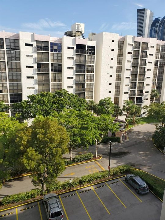 Multi-story residential building exterior featuring private balconies, expansive landscaped grounds with mature trees, and designated surface parking