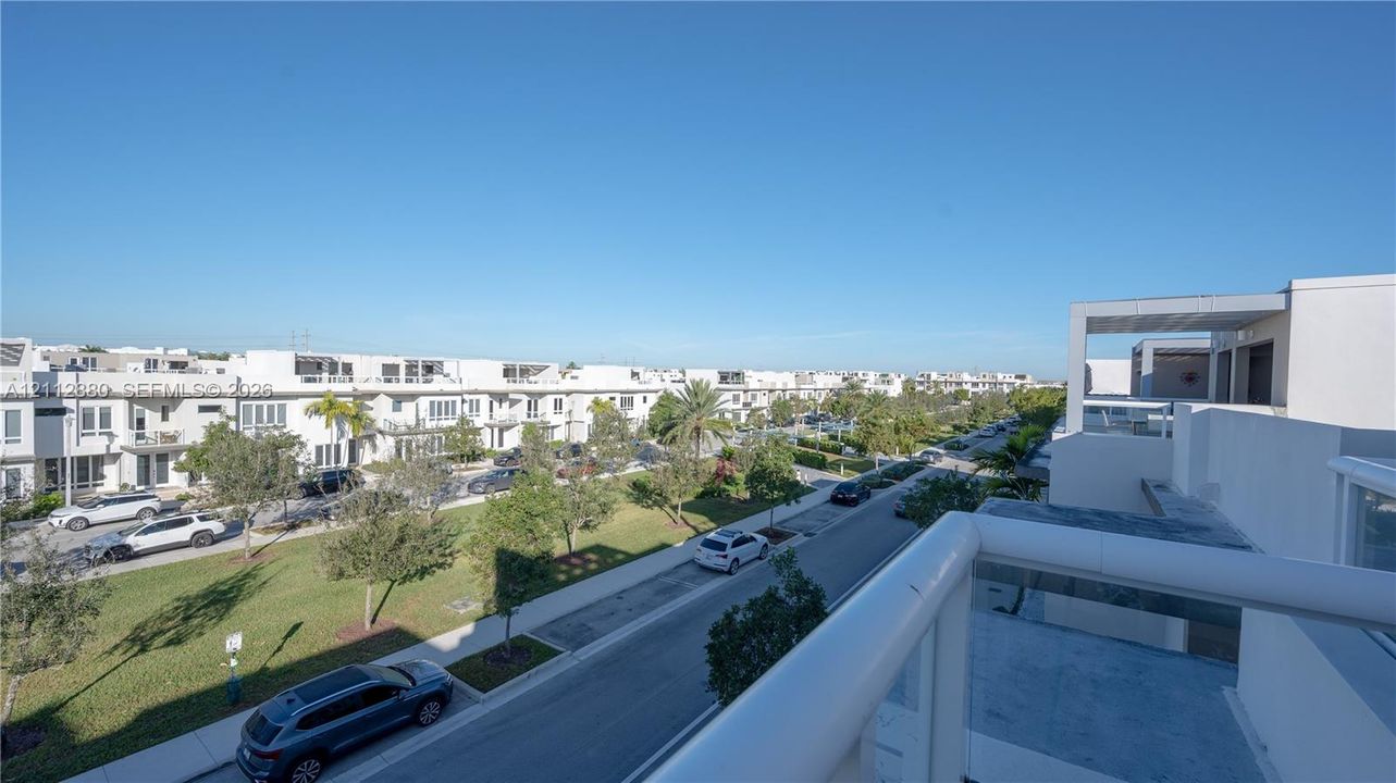 Elevated balcony featuring a white metal railing and a concrete deck surface, overlooking a paved street with green spaces and palm trees
