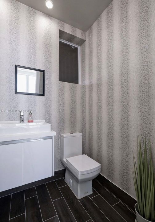 Wall-mounted white vanity with a rectangular sink, porcelain toilet, patterned wall covering, and dark wood-finish tile flooring