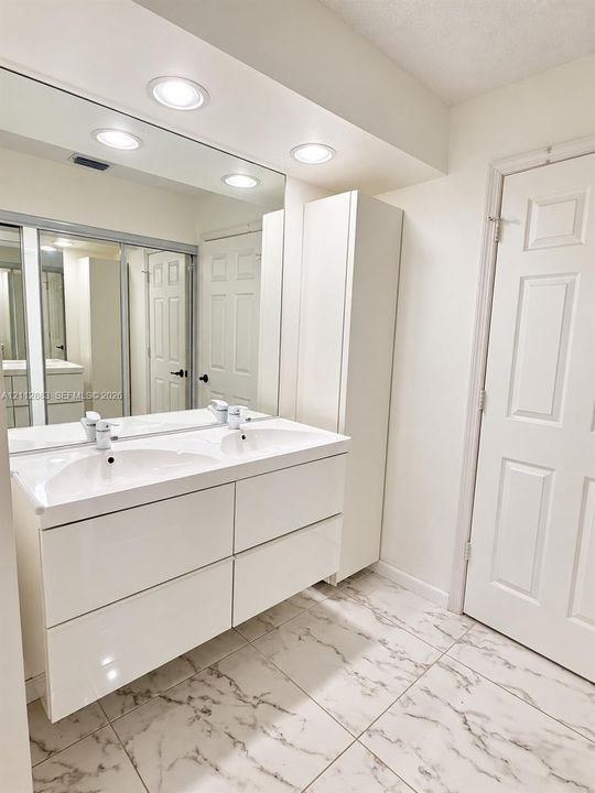 Dual-sink vanity with white high-gloss cabinetry, integrated countertop, full-width vanity mirror, built-in floor-to-ceiling storage cabinet, and large-format tile flooring with a veined pattern