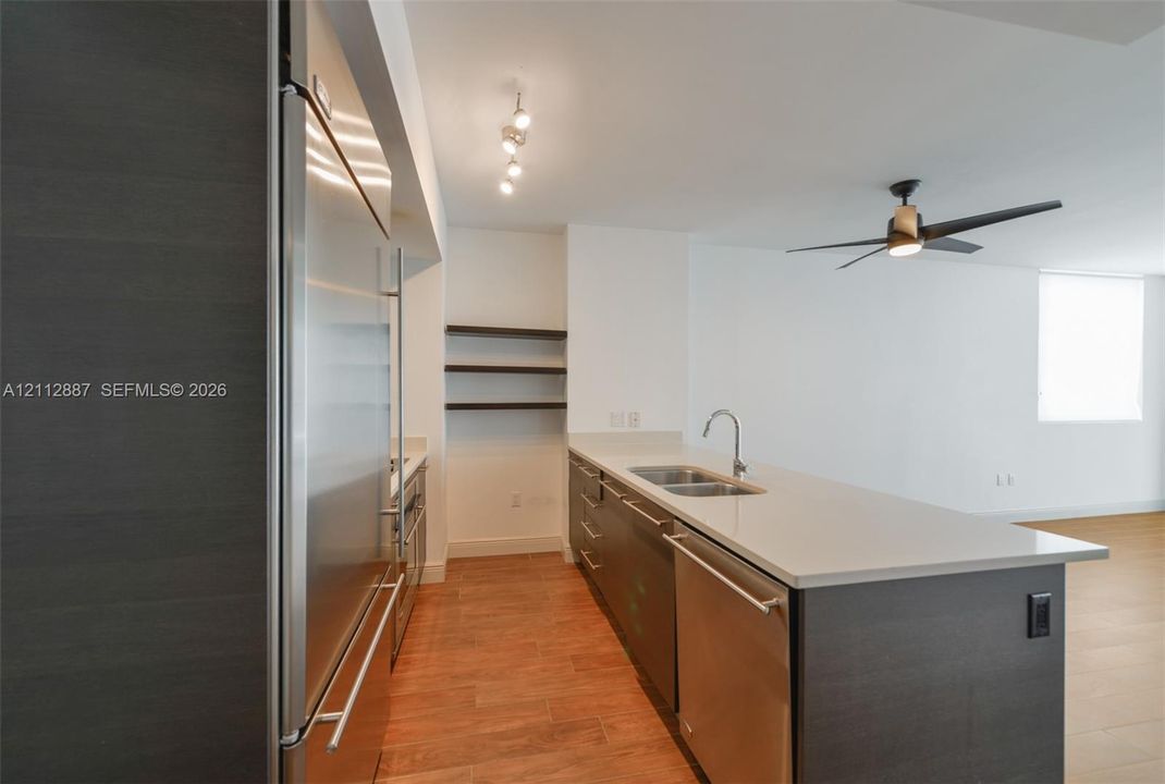 Kitchen featuring custom dark-toned cabinetry, stainless steel appliances, a stone countertop island with a dual-basin sink, wood-finish flooring, and multi-tier wall shelving