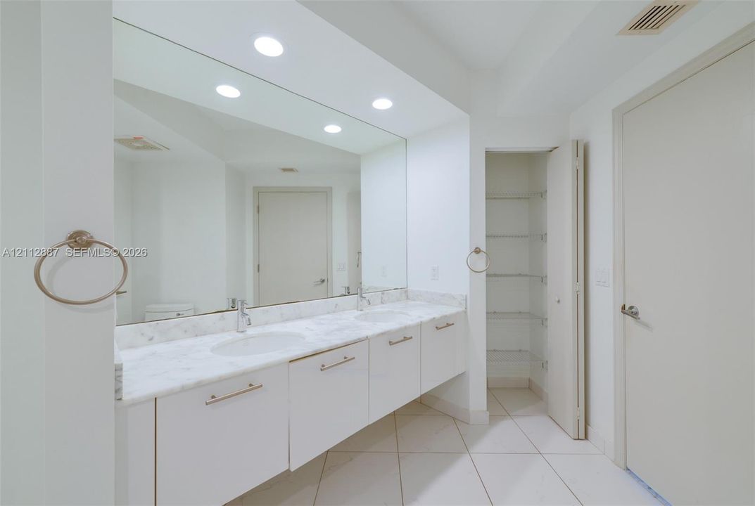 Floating vanity with a solid-surface countertop and dual integrated sinks, floor-to-ceiling vanity mirror, recessed lighting, and built-in closet shelving